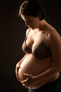 Babybauch Fotografie 5 Eine schwangere Frau steht vor dunklem Hintergrund und hält ihren Bauch sanft mit beiden Händen. Sie blickt mit gelassenem Gesichtsausdruck nach unten, trägt einen braunen BH und eine dunkle Hose und verkörpert damit die Essenz von Babybauch Fotografie. Die Beleuchtung hebt ihr Profil und ihren Babybauch wunderschön hervor.