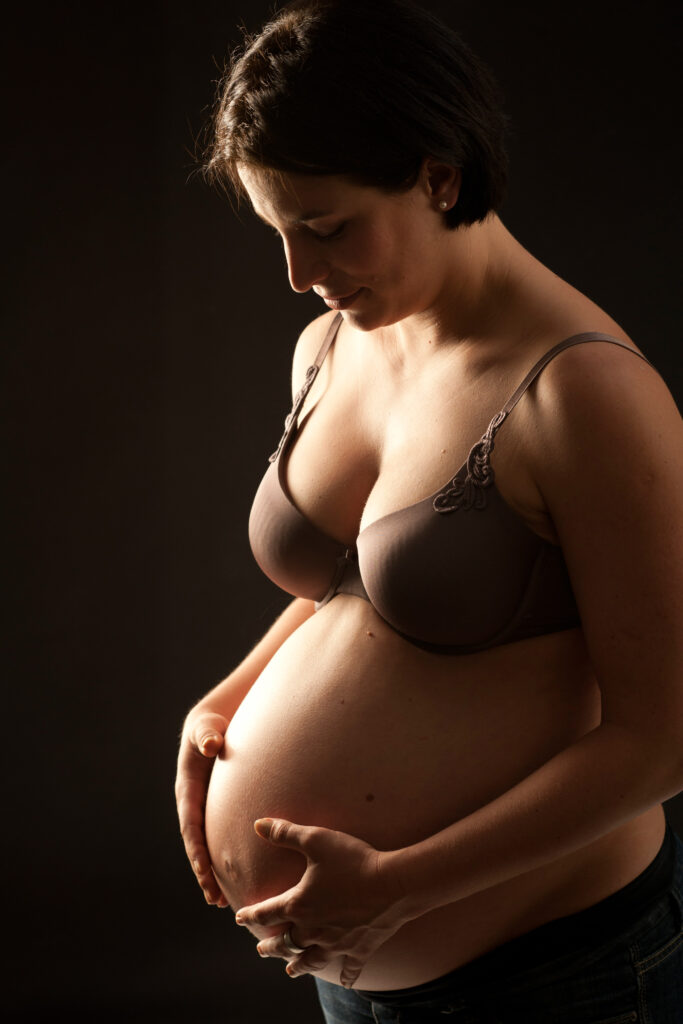 Eine schwangere Frau steht vor dunklem Hintergrund und hält ihren Bauch sanft mit beiden Händen. Sie blickt mit gelassenem Gesichtsausdruck nach unten, trägt einen braunen BH und eine dunkle Hose und verkörpert damit die Essenz von Babybauch Fotografie. Die Beleuchtung hebt ihr Profil und ihren Babybauch wunderschön hervor.